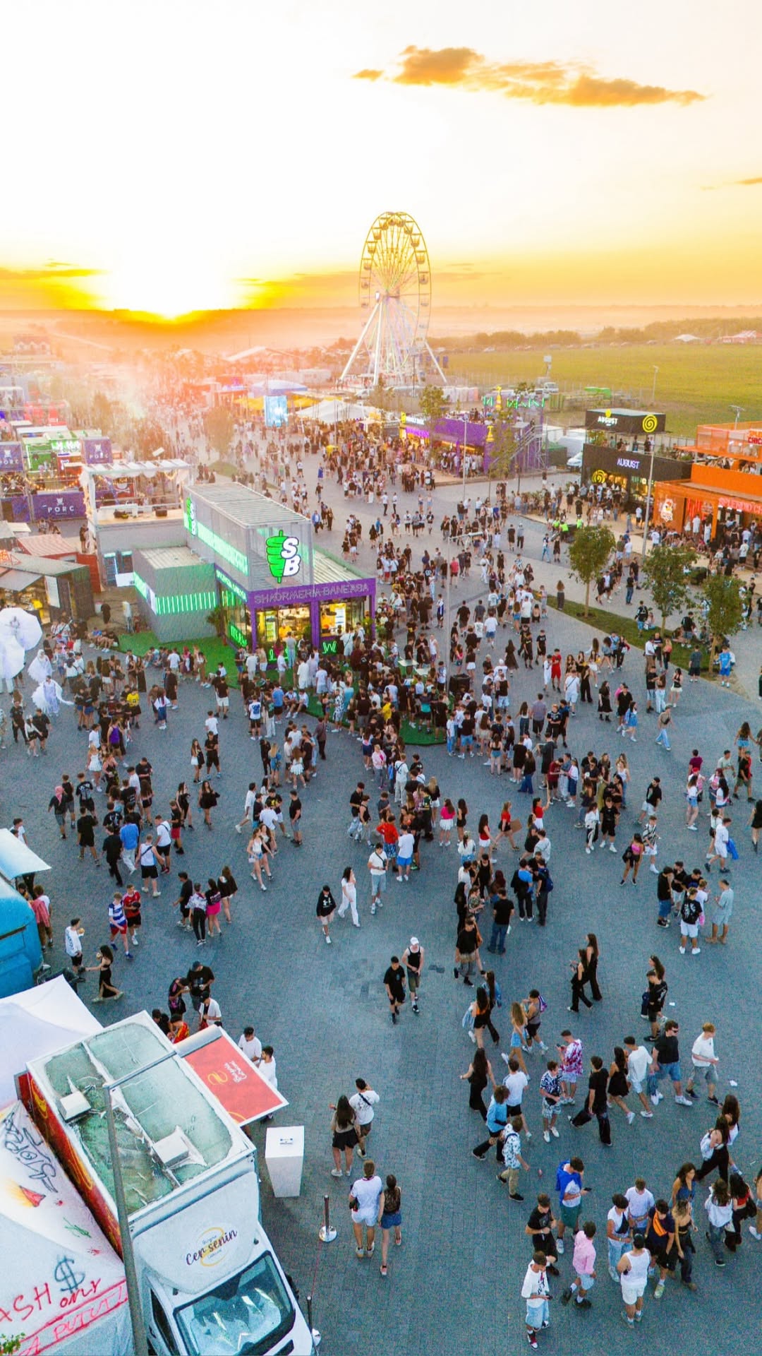 Zona festivalului la apus, cu public, standuri și roată panoramică